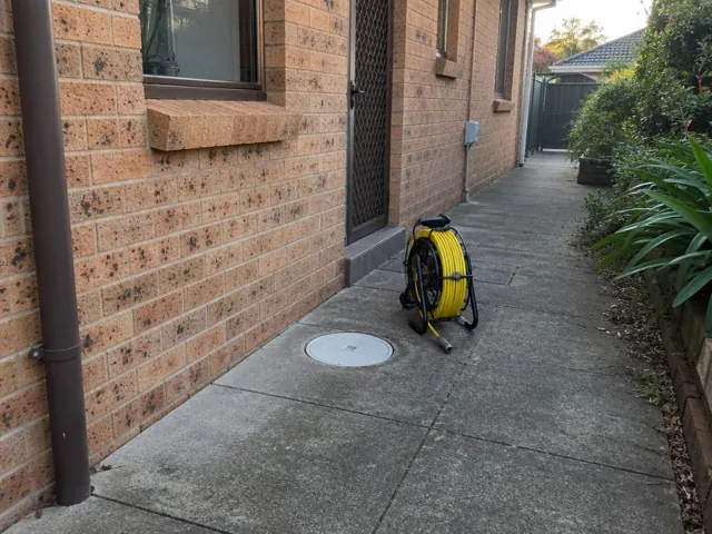 Capped drain inspection opening beside a coiled camera reel on a concrete side path by brick walls in Bonnyrigg