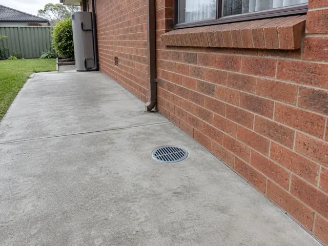Outdoor concrete gully grate beside a brick wall and lawn, with dry paved area in Bonnyrigg